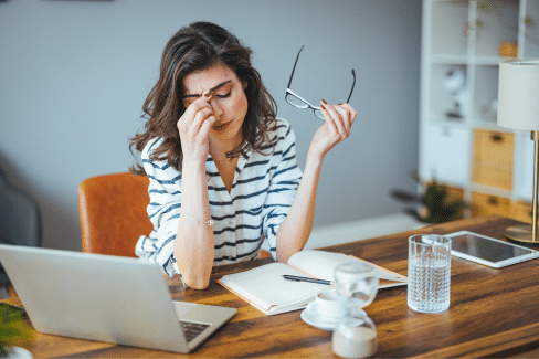 brunette woman in a striped shirt at a desk holding her glasses and rubbing the bridge of her nose.